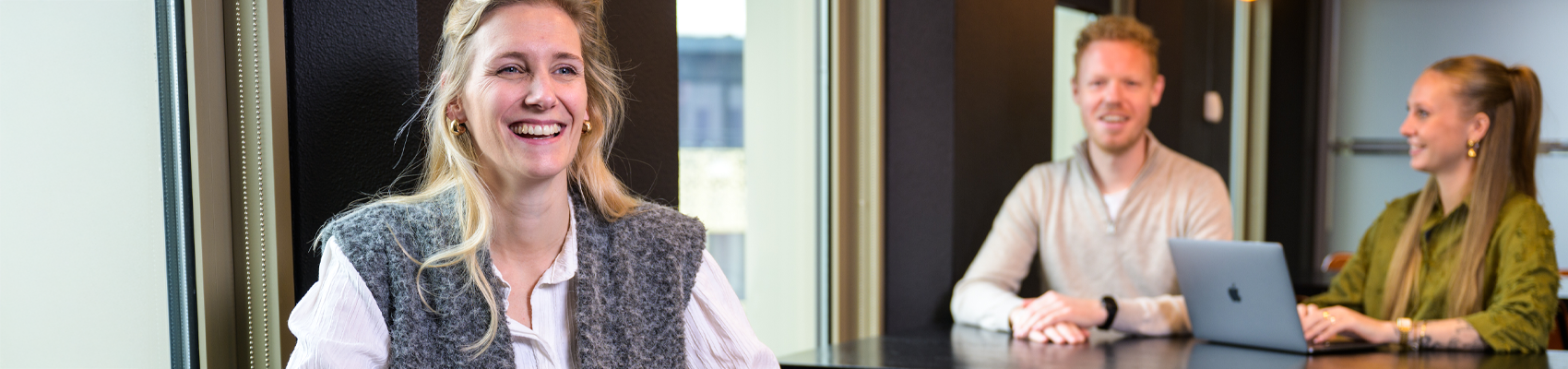 Banner 1700×400 Collega lacht en twee collega’s zijn op de achtergrond aan het werk