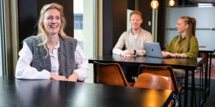 Banner 800×500 mobiel Collega lacht en twee collega’s zijn op de achtergrond aan het werk