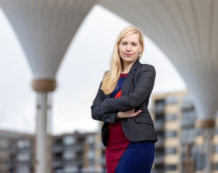 Persoon in zakelijke kleding staat met armen over elkaar onder grote betonnen bogen, met gebouwen op de achtergrond